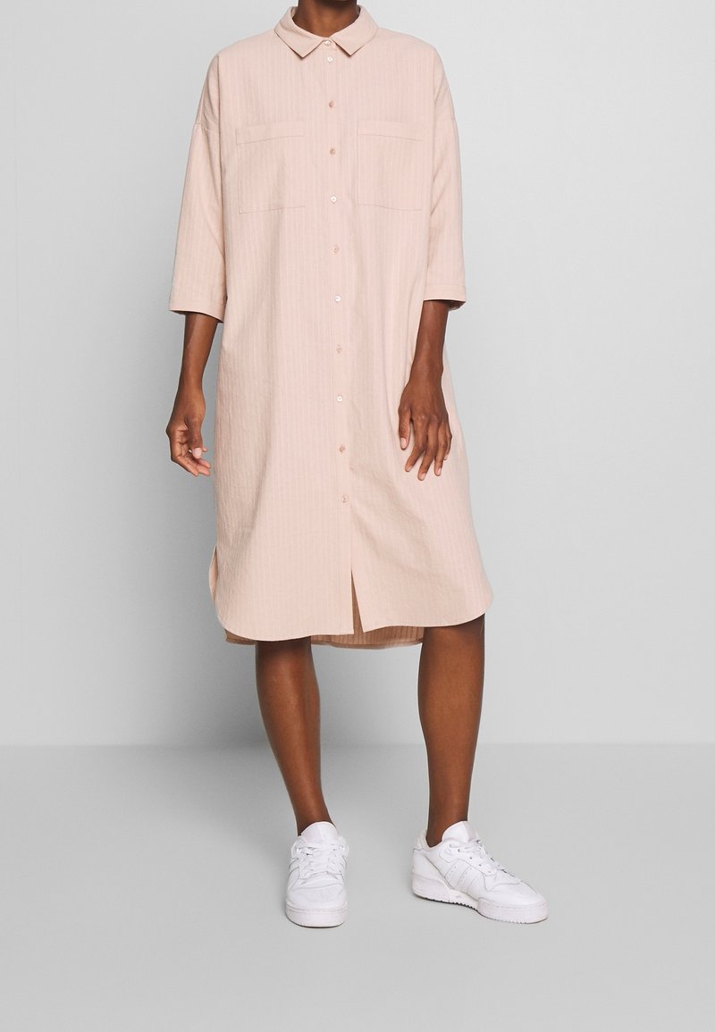 Personne portant une robe-chemise rose clair, boutonnée, longueur genou, manches trois-quarts et baskets blanches, debout devant un fond gris.