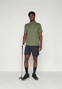 Άνδρας με ελιά πράσινο πόλο, μαύρες βερμούδες, γάντι του γκολφ και παπούτσια, κρατώντας μια ρακέτα του γκολφ, στέκεται μπροστά σε απλή ανοιχτόχρωμη φόντο.