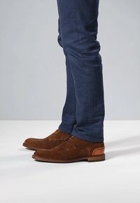 Braune Wildlederschuhe mit Fischgrätstruktur, runder Spitze und flachem Absatz, kombiniert mit dunkelblauen, eng anliegenden Hosen.