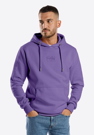 Man with short hair and beard wearing a purple hoodie with drawstrings and front pocket, hands in pockets, looking to the side.