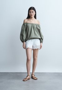 Γυναίκα που στέκεται φορώντας μια ελιά μπλούζα χωρίς ώμους, λευκά σορτς με διακοσμητικές τρύπες και καφέ σανδάλια με λουράκια μπροστά σε έναν απλό γκρι τοίχο.