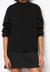 Pull noir en tricot côtelé avec un col montant et des manches raglan, présentant une coupe décontractée et un motif texturé, associé à une jupe noire.