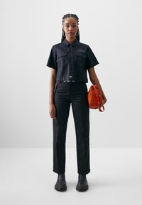 Black cropped button-up shirt with two chest pockets, paired with black straight-leg pants and black platform boots. Holding an orange bag.