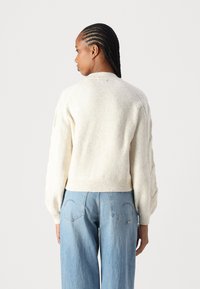 Sweater cropped na cor creme, com um acabamento de malha texturizada, bainha e punhos canelados, e mangas raglã, combinado com jeans de perna larga em azul claro.