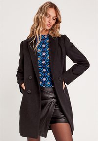 Black double-breasted wool coat with notched lapels, front pockets, and a textured finish, layered over a patterned blue top and leather skirt.