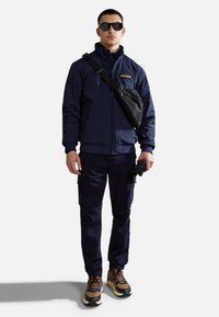 Marinfärgad bomberjacka med en svart crossbody-väska, tillsammans med mörka byxor och bruna sneakers. Inkluderar solglasögon och en texturerad tyg.