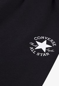 Čierna tkanina s pletenou textúrou, obsahujúca biele okrúhle logo, ktoré zahŕňa hviezdu a text "CONVERSE ALL STAR" a "Chuck Taylor."