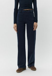 Tumši zili platām kājām džinsi ar vidēju jostas augstumu, priekšējām un aizmugurējām kabatām, kā arī gludu tekstūru. Saskaņoti ar brūnām ikdienas kurpēm.