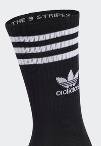 Medias de algodón negras con tres líneas blancas en la parte superior, que presentan un logo tejido de un trébol con el texto "adidas" debajo en un color contrastante.