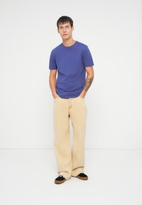 Tricou scurt cu mâneci scurte de culoare mov, asortat cu pantaloni largi bej deschis și pantofi negri tip slip-on. Modelul stă cu mâinile în buzunare.