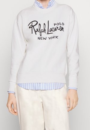 Personne portant un sweat-shirt blanc Ralph Lauren avec un texte de logo noir, superposé sur une chemise rayée bleu et blanc à boutons et un pantalon clair.