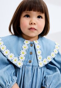 Robe en denim bleu avec un grand col orné de marguerites blanches au centre jaune ; boutons en bois et manches froncées ajoutent des détails.