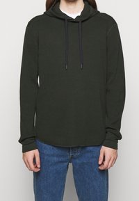 Sudadera de punto waffle verde oliva con un corte relajado, mangas largas, cordones ajustables negros y dobladillo redondeado. Combinada con jeans azul.