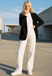 Young woman with long blonde hair wearing white pants, white sneakers, white shirt, and black blazer, standing outdoors on a concrete surface.