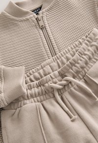 Chaqueta beige de punto con cremallera frontal y pantalones joggers a juego con cintura elástica, puños acanalados y tejido texturizado.