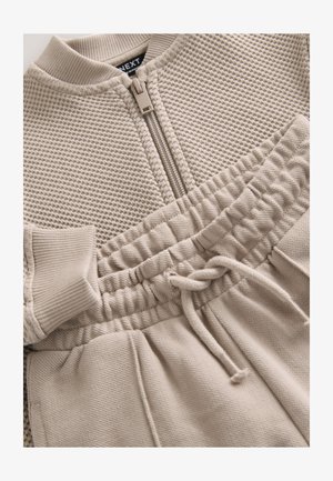 Bež pletena jakna z zadrgo spredaj in ujemajoče športne hlače z elastičnim pasom, rebrastimi manšetami in teksturirano tkanino.