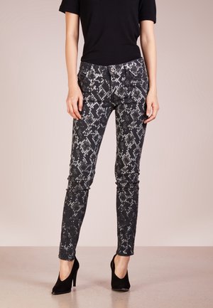 Nainen, jolla on mustat ja harmaat käärmekuvioiset kapeat housut, musta lyhythihainen paita ja mustat korkokengät, seisomassa neutraalilla lattialla.