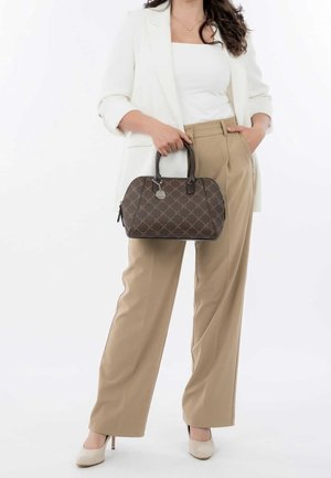 Woman in beige trousers and white blazer holding a brown patterned handbag with black handles, standing against a white background.