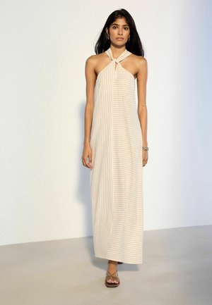 Frau mit langen dunklen Haaren trägt ein beige-weiß gestreiftes Neckholder-Maxikleid und Sandalen, steht vor einem schlichten hellen Hintergrund.