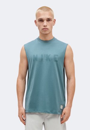 Jeune homme aux cheveux blonds courts portant un débardeur bleu Nike et un short clair, debout devant un fond uni.