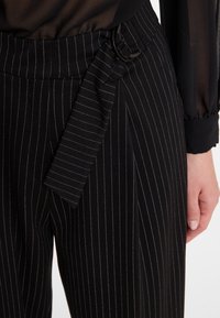 Zwarte pinstripe broek met een brede tailleband, voorzien van een riem met een ronde gesp, gestructureerde stof en gedrapeerd lintdetail.