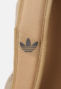 Bežový batoh s textúrovanou tkaninou a prešitými okrajmi, s čiernym logom Adidas trefoil na bočnej strane.
