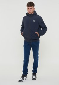 Marineblå hættejakke med lynlås foran, elastiske manchetter og snøre. Har et lille hvidt logo på brystet, kombineret med blå jeans og sneakers.