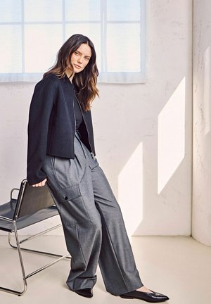Veste noire courte sur un haut noir ajusté, associée à un pantalon gris ample taille haute avec de grandes poches latérales. Mocassins noirs à bout pointu.