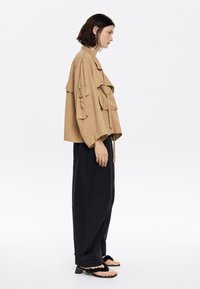 Tan cropped jack met meerdere zakken, losse zwarte trousers en zwarte sandalen met een gestructureerde band. Gladde stof, relaxte pasvorm.