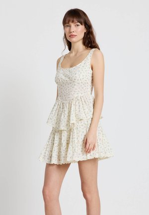 Femme portant une robe sans manches couleur crème à motifs floraux, avec une jupe à volants superposés, debout devant un fond clair uni.