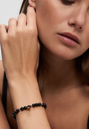 Woman wearing a black beaded bracelet with small gold accents, resting her hand near her face against a neutral background.
