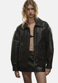 Chaqueta de cuero negra con un gran cuello, cremallera frontal, dos bolsillos y un corte relajado, combinada con una falda de cuero a juego.
