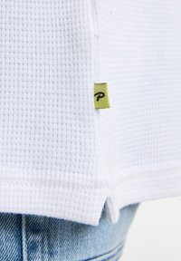 Vit tröja i våfflad stickning med en grön etikett med ett svart "P" längst ner på sömmen, uppvisad mot ljusblå denim.