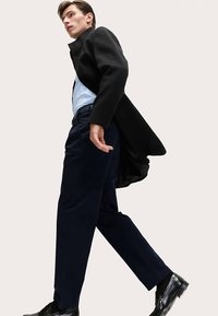 Schwarzer Mantel, hellblaues Hemd, dunkle Hose, glänzende schwarze Schuhe; Modell läuft mit flatterndem Mantel vor neutralem Hintergrund.