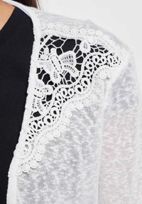Weiße Spitzenstrickjacke mit detaillierter floraler Spitzenborte an der Schulter, aus leichtem, strukturiertem Stoff gegen ein dunkleres Oberteil.