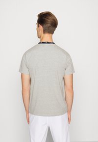 Młody mężczyzna z krótkimi brązowymi włosami stoi przodem do białej ściany, ubrany w jasnoszary t-shirt i białe spodnie.