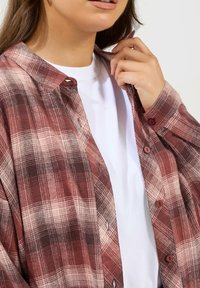 Czerwono-beżowa koszula flanelowa w kratę z zapinanym kołnierzykiem, noszona na białym t-shircie. Materiał wydaje się miękki o teksturowanym wykończeniu.