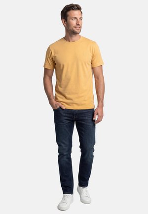 Mand stående iført en sennepsgul t-shirt, mørkeblå jeans og hvide sneakers, med den ene hånd i lommen og kigger til siden.