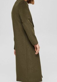 Kobieta w długim, oliwkowozielonym wełnianym płaszczu, stojąca bokiem na jednolitym jasnoszarym tle.