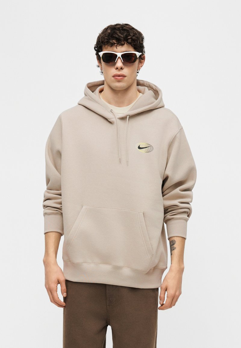 Uomo che indossa una felpa con cappuccio beige Nike con tasca frontale e logo, pantaloni marroni, occhiali da sole con montatura bianca e un tatuaggio piccolo sul polso sinistro.