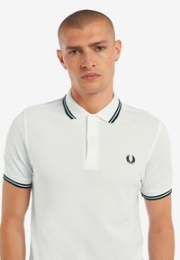 Witte polo met een zwartgestreepte kraag en manchetten, voorzien van een klein geborduurd logo aan de linkerkant. Soepele katoentextuur.