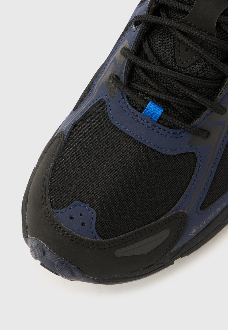 Black and navy athletic shoe with a mesh upper, textured surface, blue accent, and reinforcing overlays. Features black laces and cushioning.