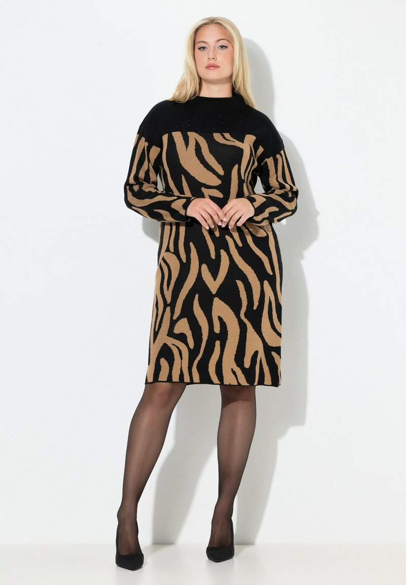 Femme blonde debout, portant une robe à manches longues à imprimé animal noir et marron, des collants noirs transparents et des talons noirs, sur fond blanc.