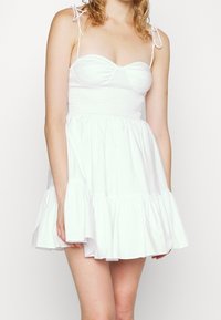 Bílá bavlněná mini šaty s nařaseným korzetem, tenkými nastavitelnými ramínky a volánkovou sukní s vrstvami. Hladká textura a neformální design.