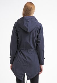 Persoon met steil rood haar in een donker marineblauwe capuchonjas met een visstaartzoom, van achteren getoond tegen een witte achtergrond.