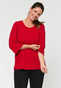 Rode textuur top met een golvend stofpatroon, ronde halslijn en driekwart mouwen, gecombineerd met zwarte broek.