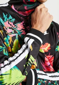 Blommig zipjacka i svart med livliga rosa, gröna och gula mönster. Ribbstickade svarta muddar med vita ränder. Slät tygtextur.