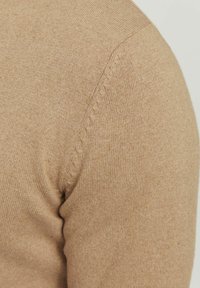 Détail de l'épaule d'un pull en tricot beige montrant une fine texture côtelée et une couture décorative subtile le long de la partie supérieure de la manche.
