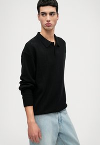 Pullover nero a maglia con collo a camicia, vestibilità comoda e polsini a coste, abbinato a jeans in denim blu chiaro. Texture morbida.