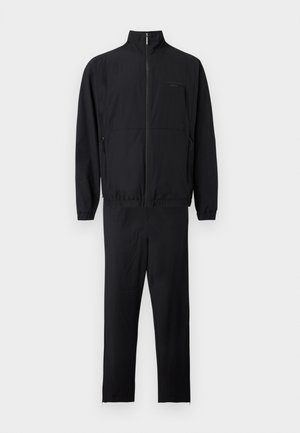 Μαύρο μπουφάν με φερμουάρ μέχρι επάνω και όρθιο γιακά, και παντελόνι με ίσια γραμμή, και τα δύο με ελαστικές μανσέτες και διακριτικές τσέπες με φερμουάρ.